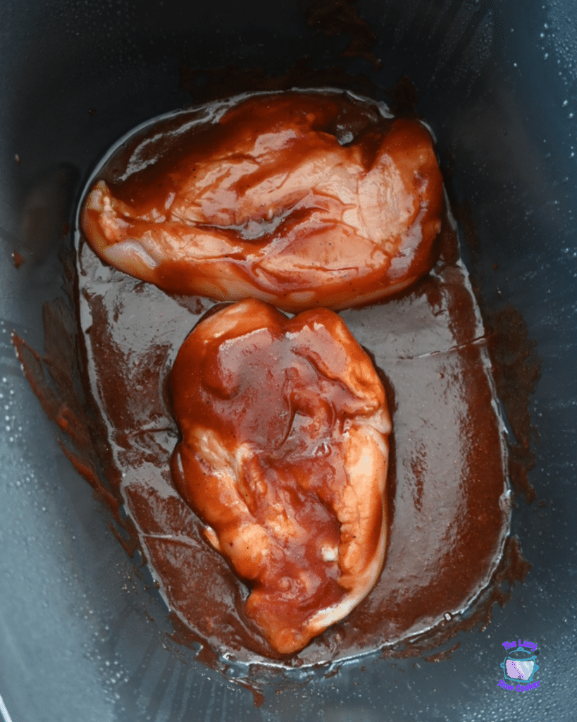 Two raw chicken breasts covered in reddish-brown BBQ sauce sit at the bottom of a slow cooker before cooking for shredded BBQ chicken sliders.