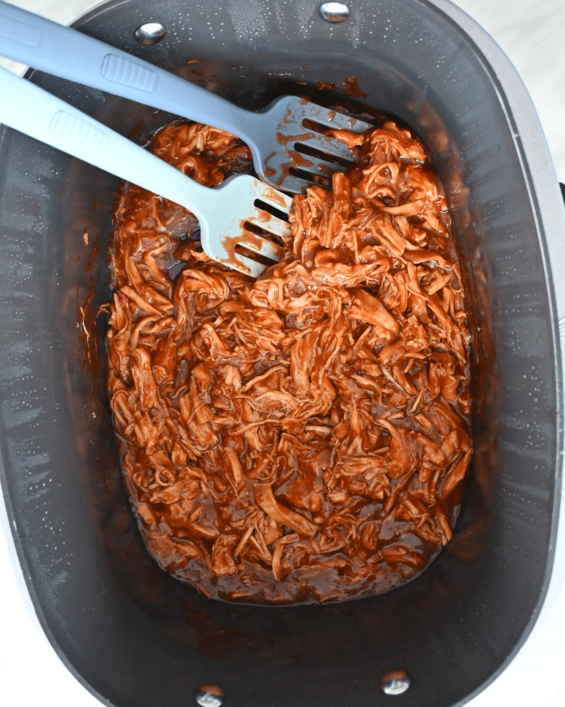 Shredded BBQ chicken sits in a black slow cooker with two blue silicone meat claws beside it, covered in a thick reddish-brown sauce.