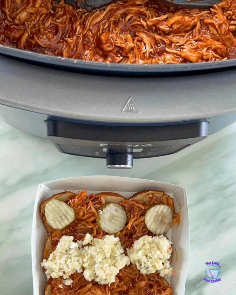 Shredded BBQ chicken in a slow cooker above sliders topped with chicken, pickles, and coleslaw in a white dish on marble.