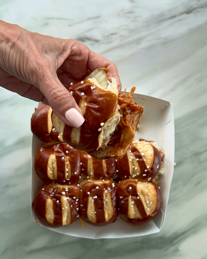 A hand with light pink nails holds a BBQ chicken slider on a pretzel bun over a tray of five more sliders on a marble countertop.