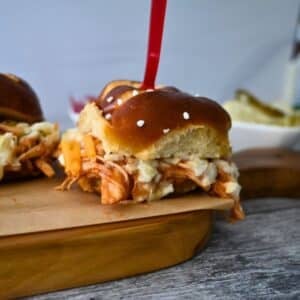 A BBQ chicken slider with coleslaw and cheese on a pretzel bun sits on a wooden board, with chips and dip behind it.