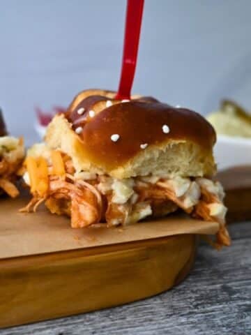A BBQ chicken slider with coleslaw and cheese on a pretzel bun sits on a wooden board, with chips and dip behind it.