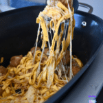 Tongs lift cheesy French Onion Meatball Pasta from a black slow cooker, melted cheese stretching as noodles are pulled up.