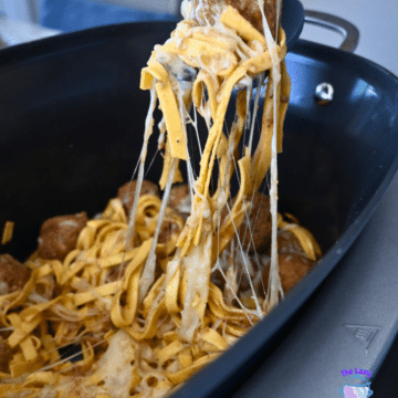 Tongs lift cheesy French Onion Meatball Pasta from a black slow cooker, melted cheese stretching as noodles are pulled up.