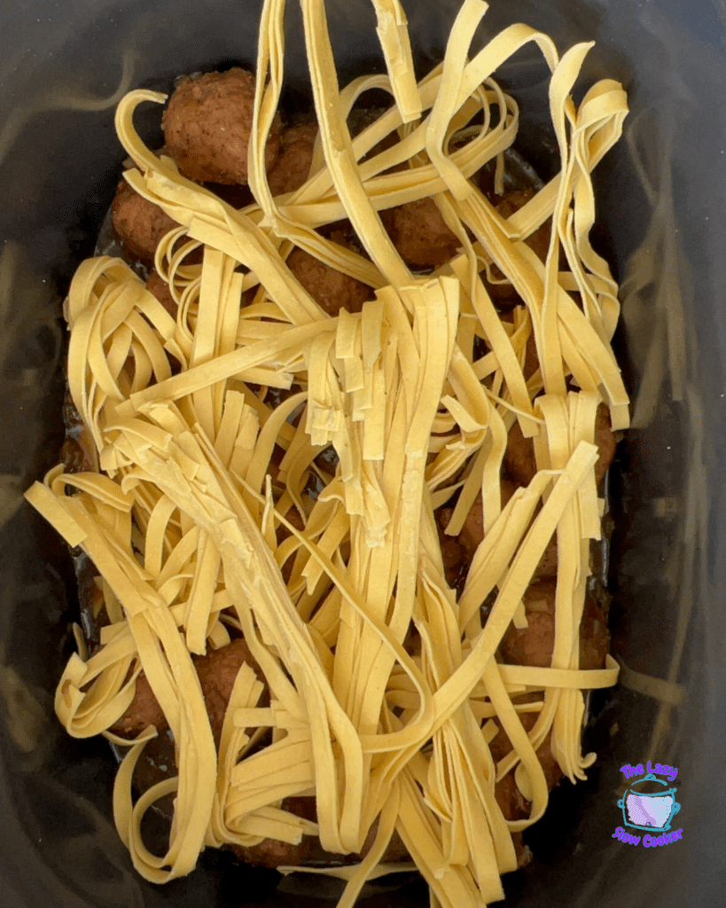 Uncooked fettuccine strips rest atop French Onion Meatball Pasta in a slow cooker, with The Lazy Slow Cooker logo in the corner.