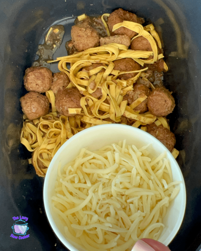 French Onion Meatball Pasta cooks in a slow cooker; a hand hovers a bowl of shredded cheese above, about to sprinkle it in.