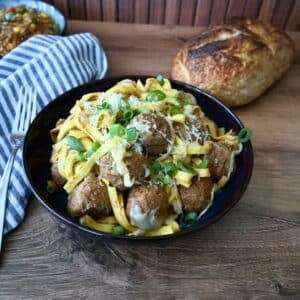 French Onion Meatball Pasta in a black bowl, topped with cheese and green onions, sits on a wooden table by bread, napkin, and fork.