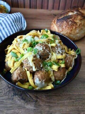 French Onion Meatball Pasta in a black bowl, topped with cheese and green onions, sits on a wooden table by bread, napkin, and fork.