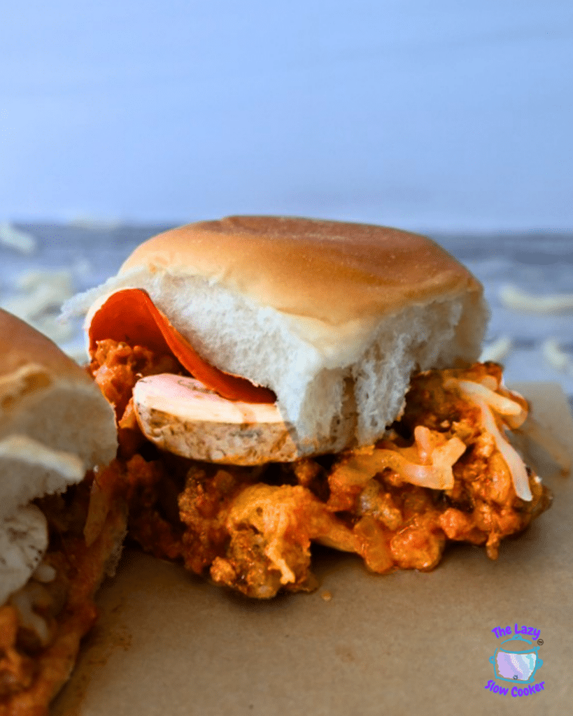 A Slow Cooker Pizza Slider with a soft bun, meat sauce, mushrooms, cheese, and pepperoni rests on a brown surface; logo in corner.