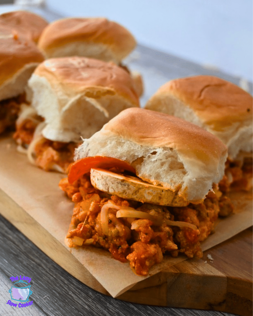 Three slow cooker pizza sliders with pepperoni sit on a wooden board; one is bitten to reveal the cheesy, meaty filling inside.
