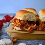 Three sliders with golden buns and hearty filling rest on a wooden board, with mushrooms and red pepper nearby. Slow Cooker Pizza Sliders.