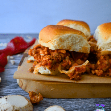 Three sliders with golden buns and hearty filling rest on a wooden board, with mushrooms and red pepper nearby. Slow Cooker Pizza Sliders.