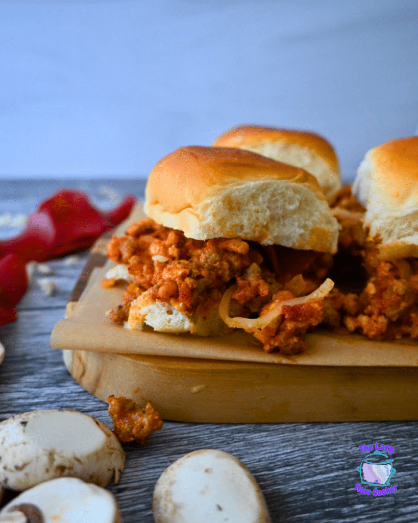 Three sliders with golden buns and hearty filling rest on a wooden board, with mushrooms and red pepper nearby. Slow Cooker Pizza Sliders.