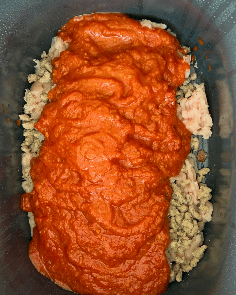 Inside a slow cooker, ground turkey, sausage crumbles ad seasonings are covered in tomato sauce, ready to cook as Slow Cooker Pizza Sliders.