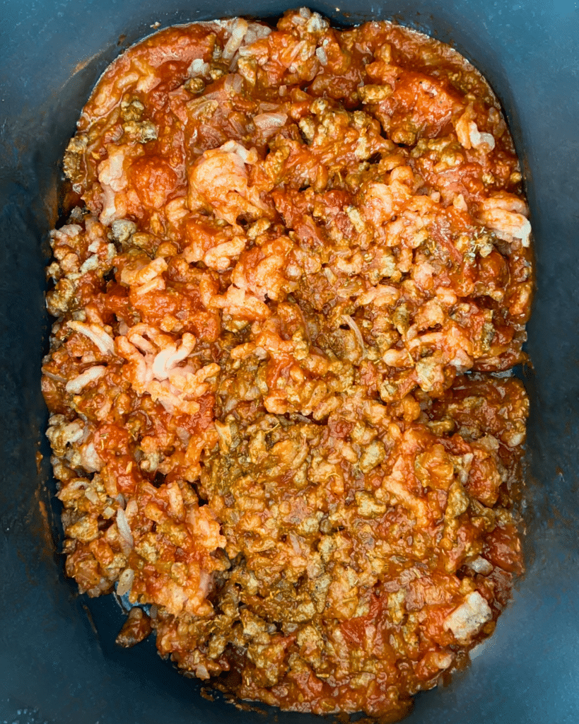 A slow cooker filled with chunky ground meat, tomato sauce, showing a red mixture with visible pieces of meat and sausage.