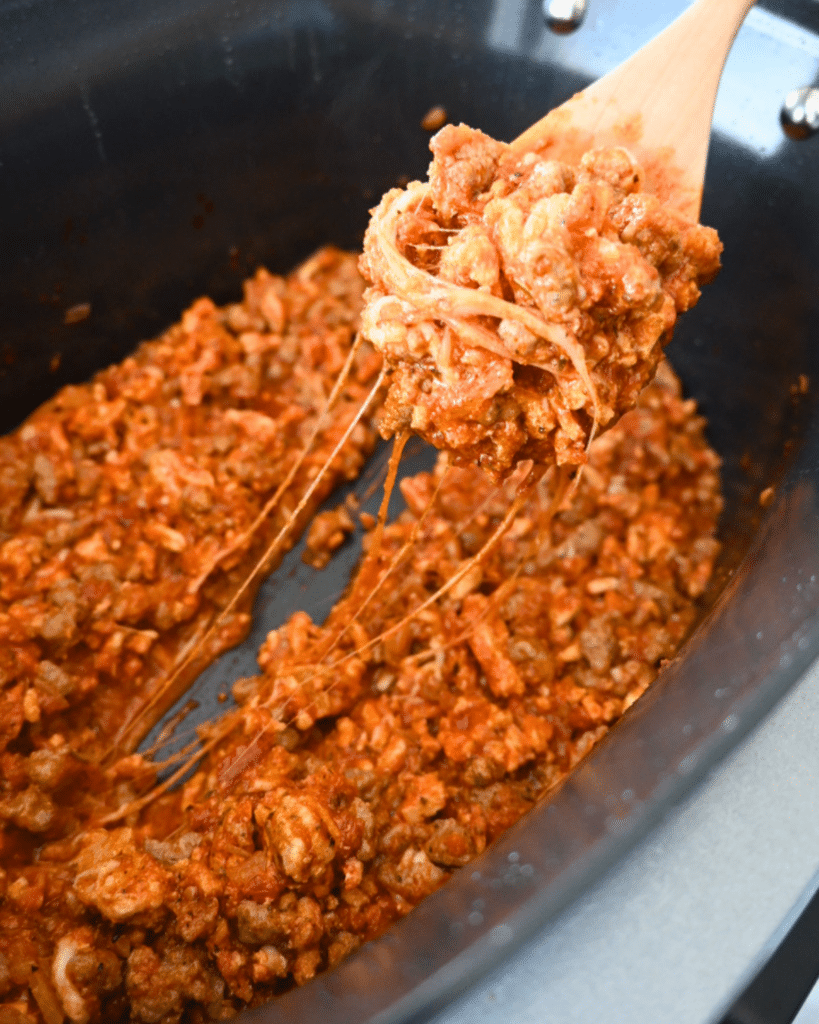 A wooden spoon lifts cheesy ground meat in tomato sauce from a black pot, with melted cheese stretching-made for pizza sliders.