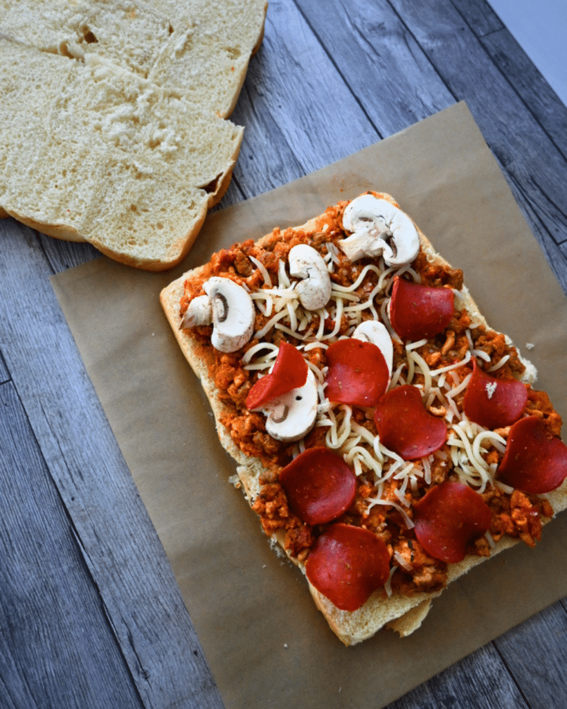 An open-faced Slow Cooker Pizza Slider with toppings rests on parchment; the top half of the bread sits nearby on a wooden surface.