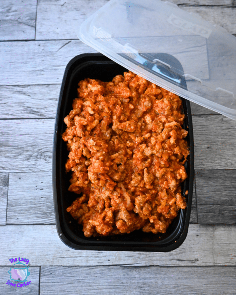 A black plastic container with its lid partly open holds leftover saucy cooked ground meat for Slow Cooker Pizza Sliders on a wooden table.