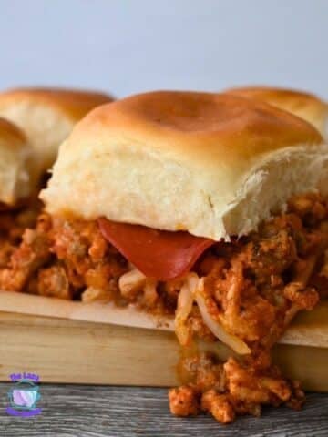 Four pizza sliders with saucy meat, melted cheese, and pepperoni on soft buns sit on a wooden board. A small logo is in the corner.