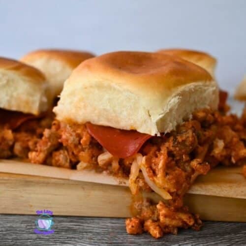 Four pizza sliders with saucy meat, melted cheese, and pepperoni on soft buns sit on a wooden board. A small logo is in the corner.