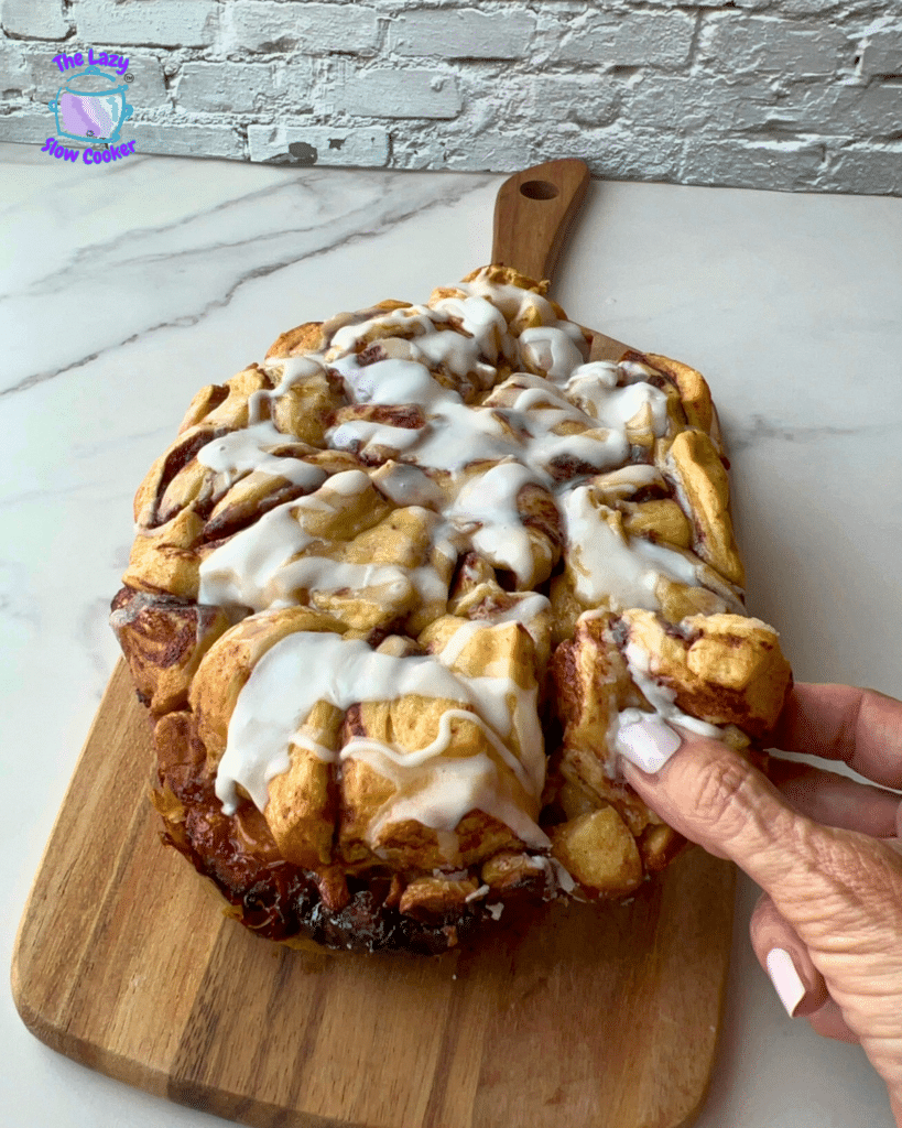 A hand pulls a soft, iced piece from a large cinnamon roll bread on a wooden board. The bread resembles slow cooker monkey bread in texture. The board rests on a marble countertop with a white brick wall behind it.
