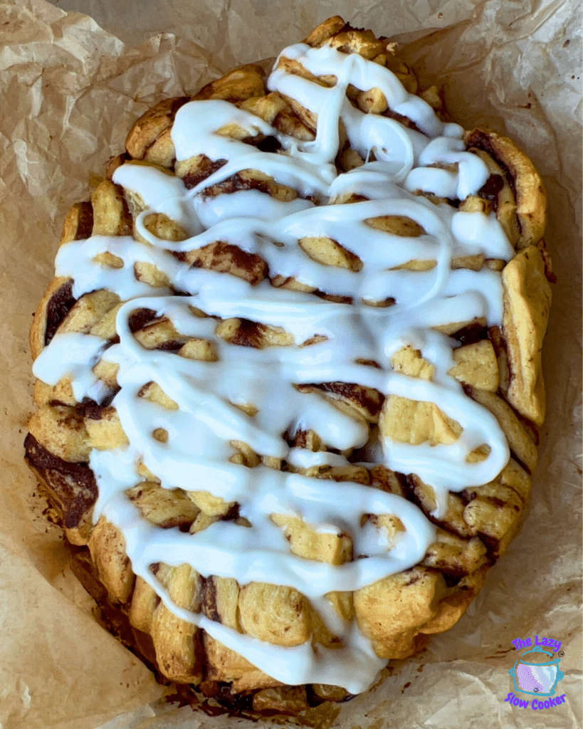 A loaf made of cinnamon rolls is topped with white icing and rests on wrinkled brown parchment paper, similar in appearance to slow cooker apple monkey bread.