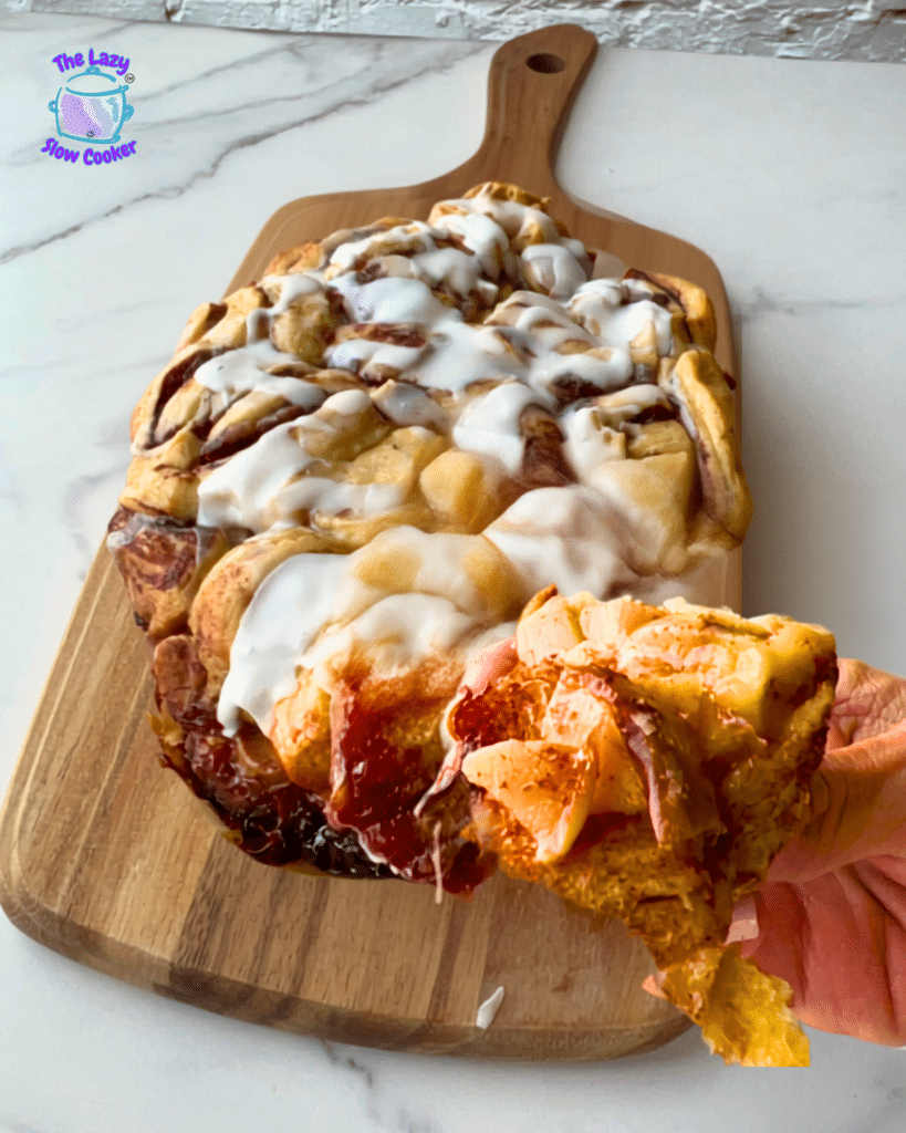A hand lifts a sticky, icing-covered slice of cinnamon roll above a wooden board filled with more frosted rolls. The Lazy Slow Cooker logo appears in the top left corner.