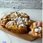 A slow cooker cinnamon roll casserole with white icing rests on a wooden board. A hand lifts out a gooey piece, showing soft layers inside. The scene is set against a white brick wall and marble countertop, with a slow cooker logo in the bottom left corner.