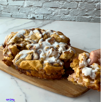 A slow cooker cinnamon roll casserole with white icing rests on a wooden board. A hand lifts out a gooey piece, showing soft layers inside. The scene is set against a white brick wall and marble countertop, with a slow cooker logo in the bottom left corner.