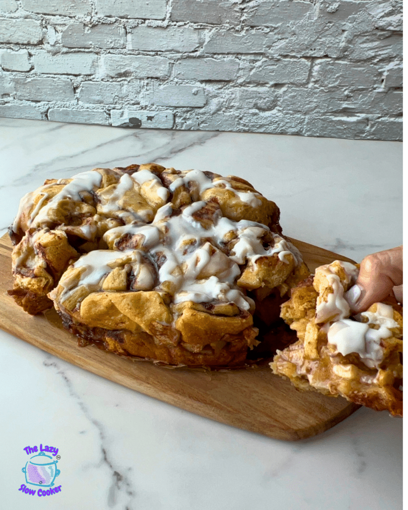 A slow cooker cinnamon roll casserole with white icing rests on a wooden board. A hand lifts out a gooey piece, showing soft layers inside. The scene is set against a white brick wall and marble countertop, with a slow cooker logo in the bottom left corner.
