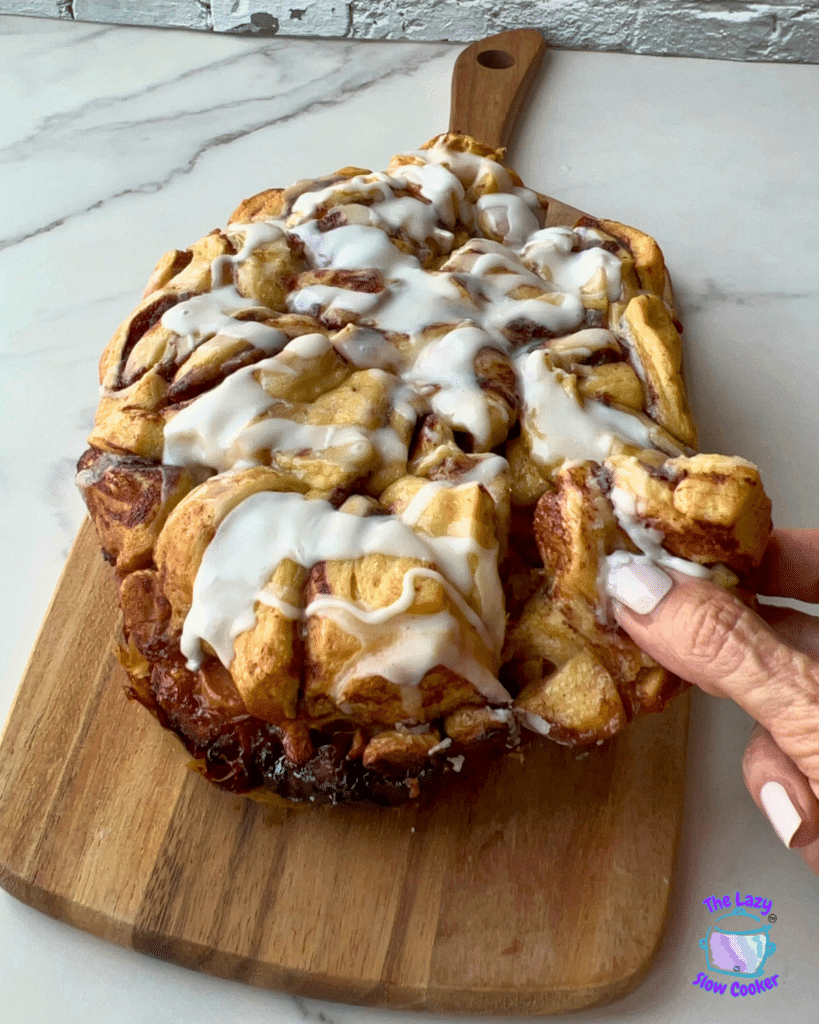 A hand tears off a piece of cinnamon roll from a loaf covered in white icing, resting on a wooden cutting board on a pale countertop. The rolls look gooey and soft, similar to the texture of slow cooker apple monkey bread.