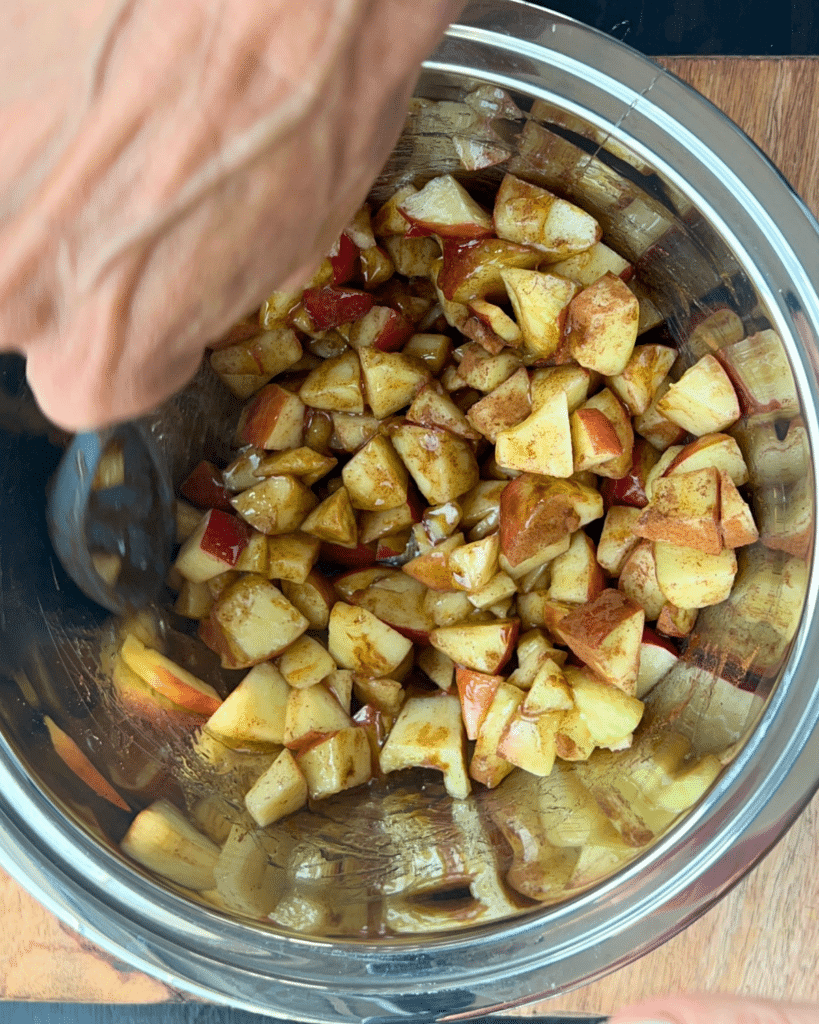 A hand mixes chopped apples dusted with cinnamon in a stainless steel bowl, getting the filling ready for slow cooker apple monkey bread.