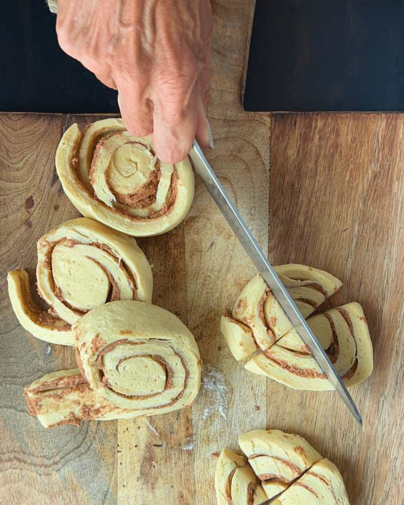 A hand cuts rolled cinnamon dough into pieces on a wooden cutting board, with several unbaked cinnamon rolls nearby, preparing ingredients for slow cooker apple monkey bread.