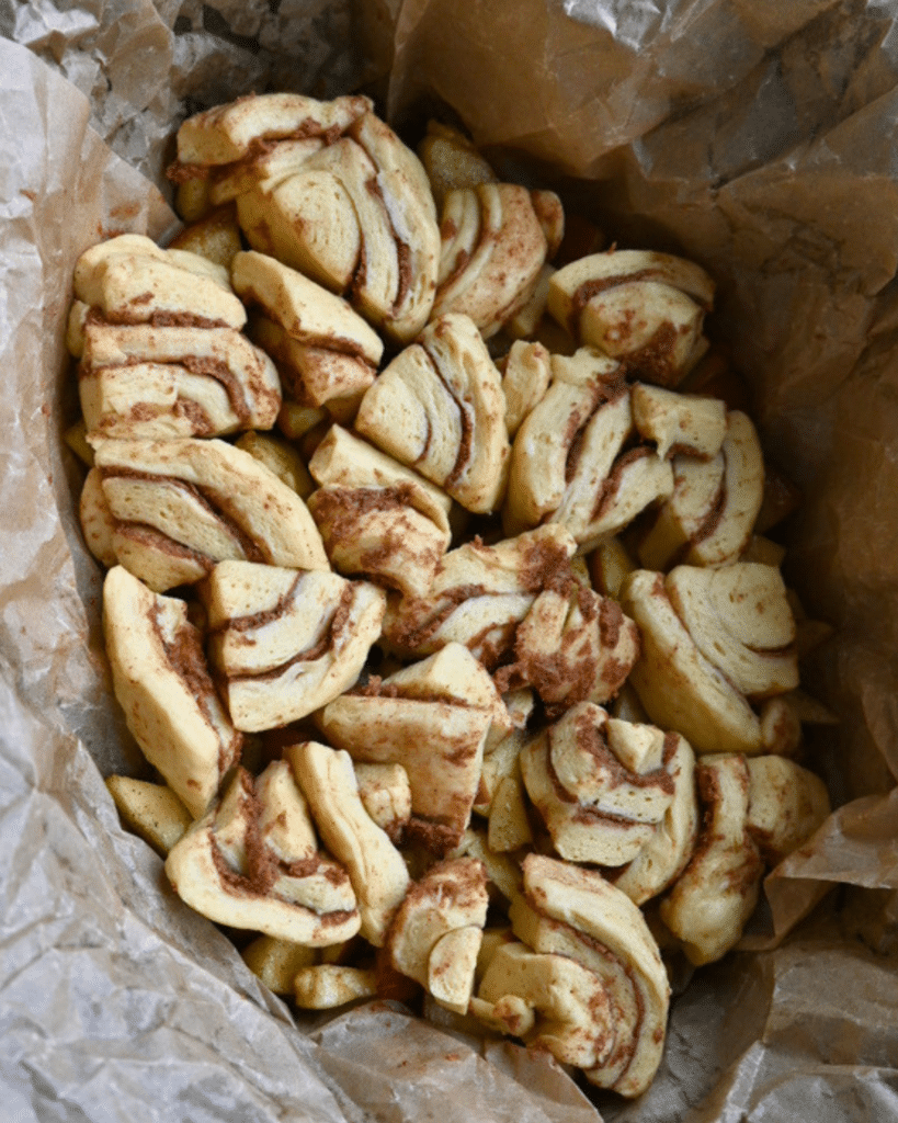 Unbaked pieces of cinnamon roll dough with clear cinnamon swirls sit on wrinkled brown parchment paper, prepared for slow cooker apple monkey bread.