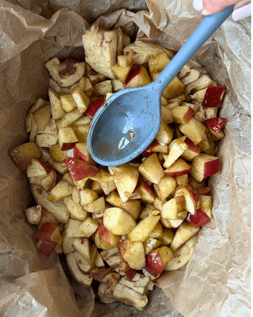 A blue spoon stirs chopped apples coated with cinnamon and spices as a second layer on top of uncooked cinnamon roll dough for slow cooker apple monkey bread.