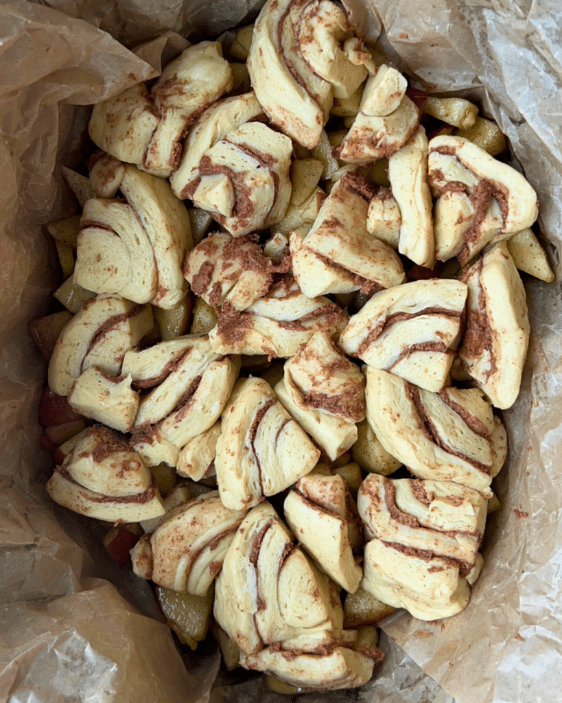 A parchment-lined baking dish holds chopped apples topped with pieces of cinnamon roll dough, ready to be baked as slow cooker apple monkey bread. The unbaked layers show swirls of cinnamon and scattered soft pastry.