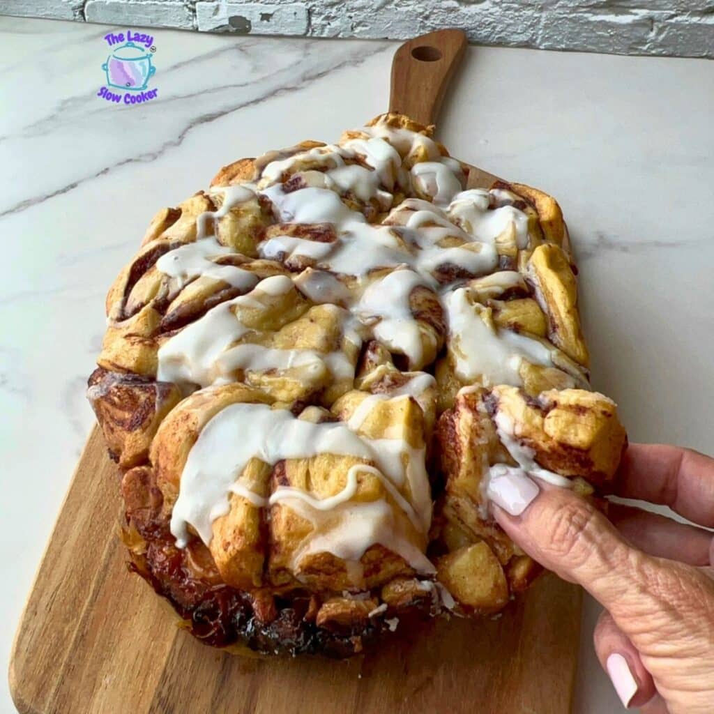 A hand pulls apart a piece of a large iced cinnamon roll, covered in white icing and resting on a wooden board atop a marble counter. The Lazy Slow Cooker logo appears in the corner.