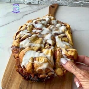 A hand pulls apart a piece of a large iced cinnamon roll, covered in white icing and resting on a wooden board atop a marble counter. The Lazy Slow Cooker logo appears in the corner.