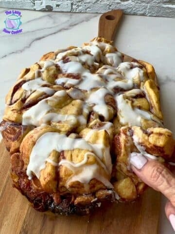 A hand pulls apart a piece of a large iced cinnamon roll, covered in white icing and resting on a wooden board atop a marble counter. The Lazy Slow Cooker logo appears in the corner.