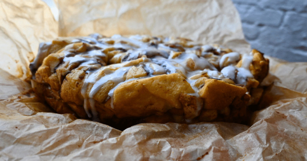 A golden-brown apple monkey bread made in a slow cooker sits on crinkled parchment paper, with white icing drizzled over the top. The bread shows soft, flaky layers and hints of spiced apple inside.
