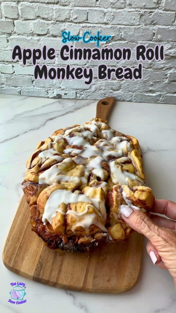 A hand lifts a piece of apple monkey bread drizzled with icing from a wooden board. The monkey bread, made in a slow cooker, sits on a marble countertop with a brick wall backdrop. Above the scene, text reads “Slow Cooker Apple Cinnamon Roll Monkey Bread.”.