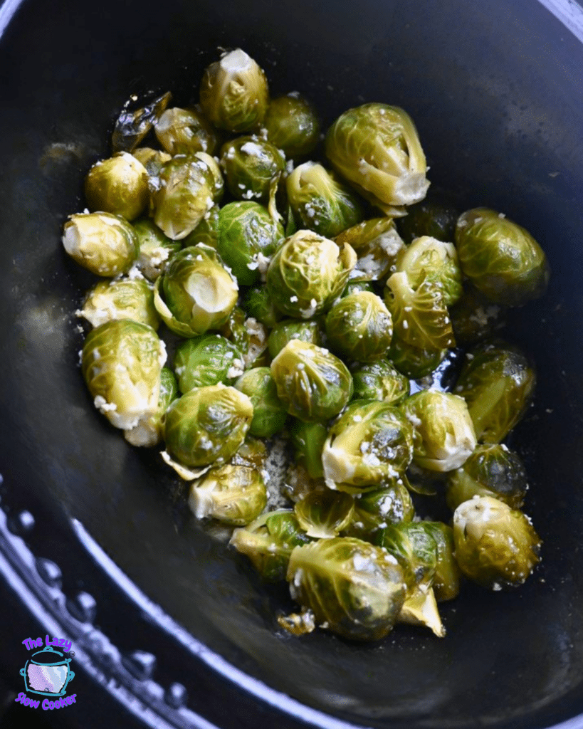 Tender, seasoned Brussels sprouts in a black slow cooker, some cut open to show texture. A small logo appears in the corner.