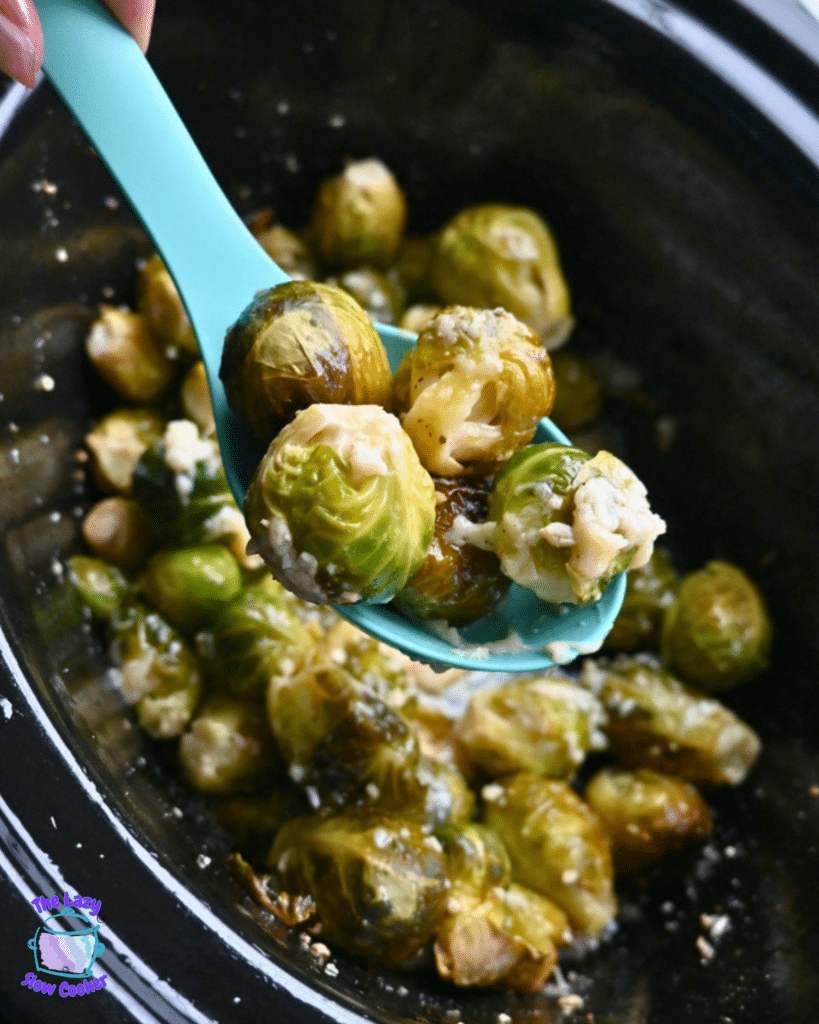 A turquoise spoon lifts cheesy, seasoned Brussels sprouts from a pot of more Brussels sprouts cooked in a slow cooker.