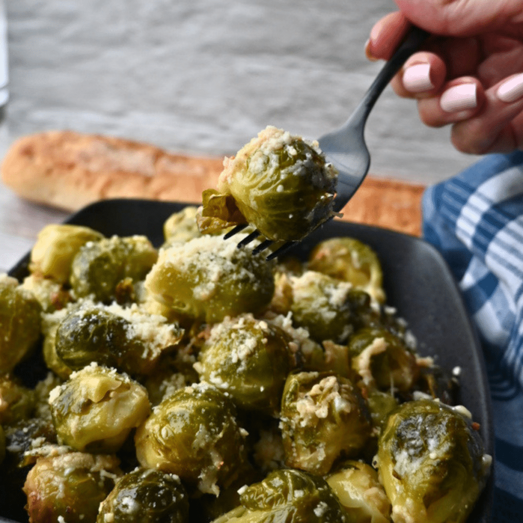 A hand holds a fork with a Brussels sprout above a plate of cheesy slow cooker Brussels sprouts, next to a baguette and blue napkin.