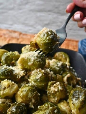 A hand lifts a Brussels sprout on a fork over a dish of cheesy slow cooker Brussels sprouts, with bread visible in the background.