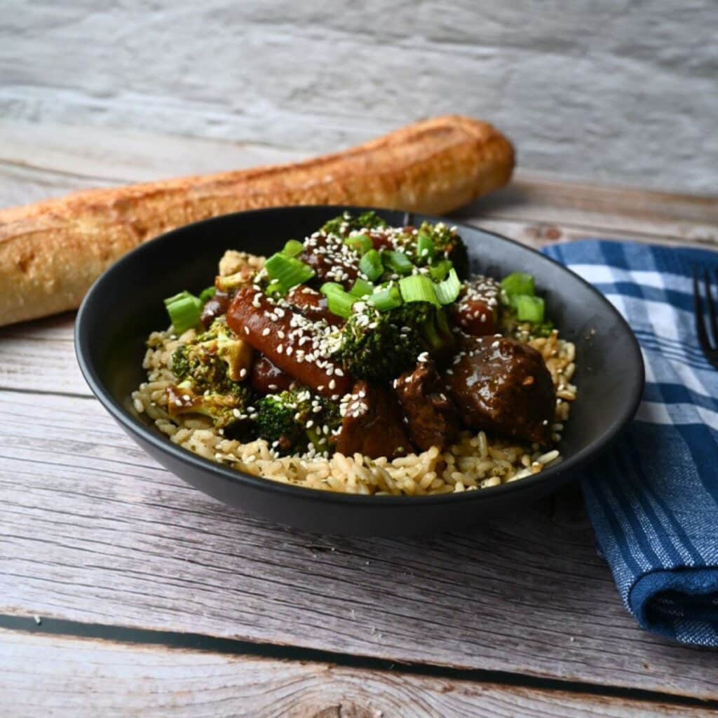 A black bowl of rice topped with slow cooker soy ginger beef, broccoli, carrots, sesame seeds, and green onions on a wooden table.