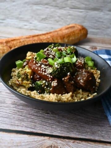 A black bowl of rice topped with slow cooker soy ginger beef, broccoli, carrots, sesame seeds, and green onions on a wooden table.
