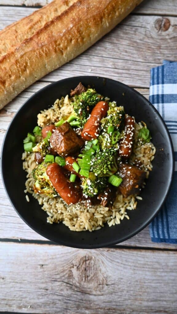 A black bowl of rice topped with slow cooker soy ginger beef, broccoli, carrots, green onions, and sesame seeds on a wooden table.