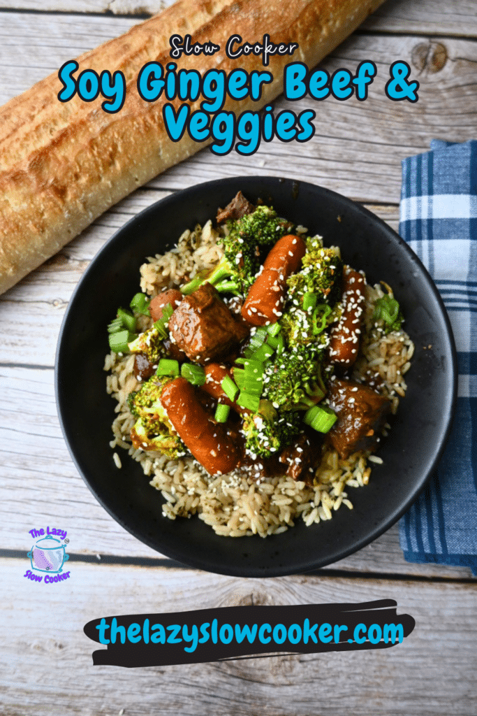 A black bowl of slow cooker soy ginger beef with carrots, broccoli, and rice sits on a wooden table by a baguette and blue plaid napkin.