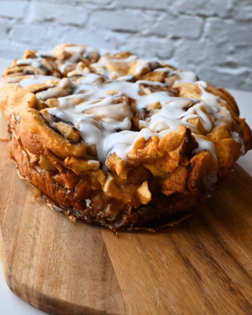 A round apple monkey bread, made in a slow cooker and topped with white icing, rests on a wooden cutting board with a light brick wall in the background.